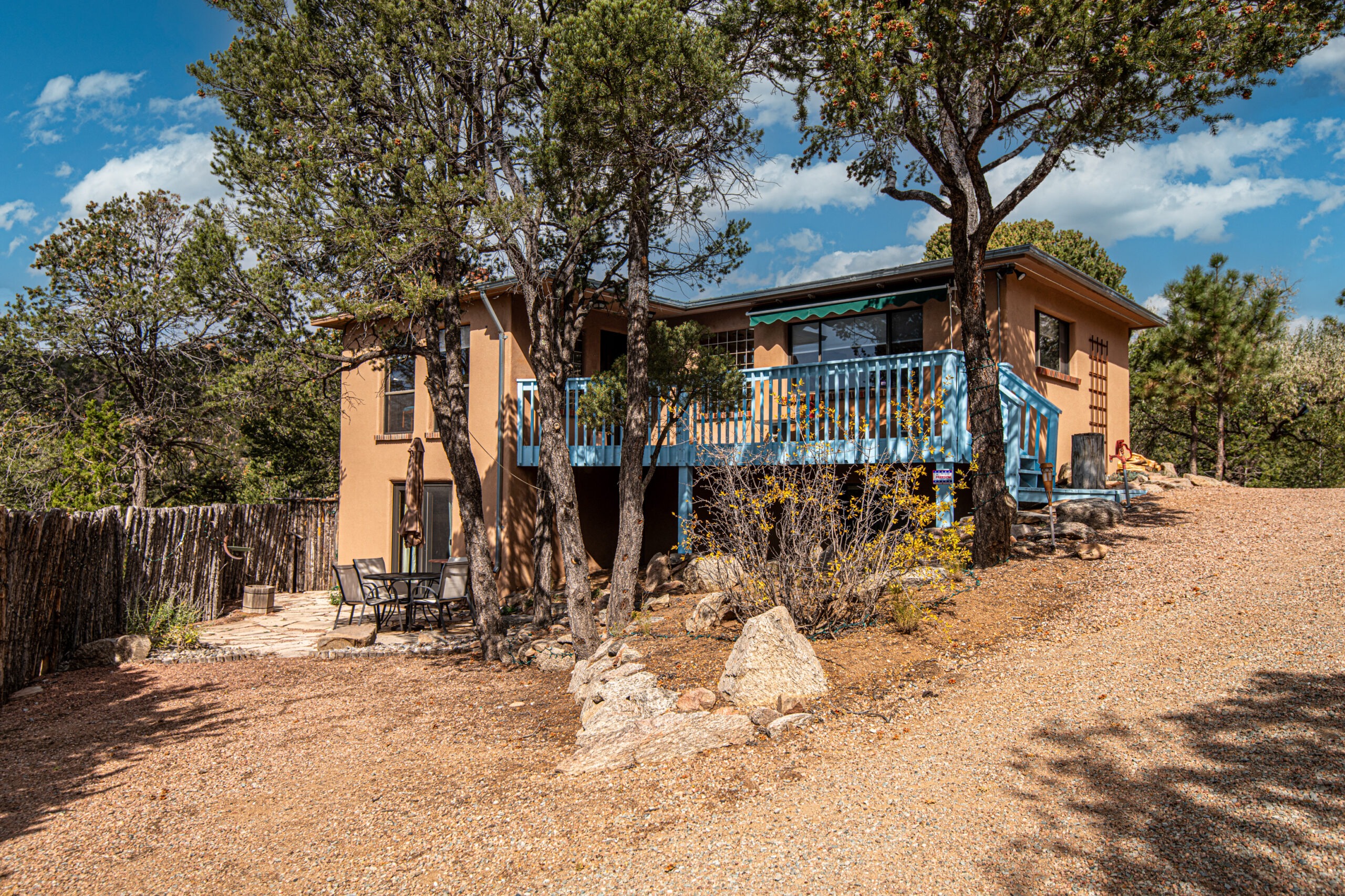 A southwestern-style stucco house with a light brown exterior and blue wooden balcony, surrounded by tall pine trees